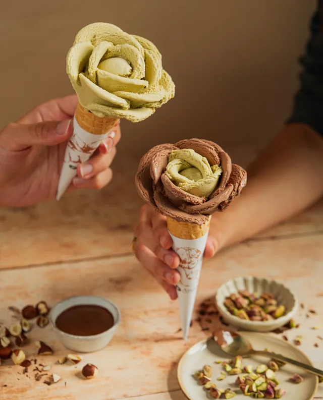 Dos conos de gelato Amorino servidos en forma de rosa, uno de pistacho y otro combinando chocolate y pistacho, sostenidos en la mano