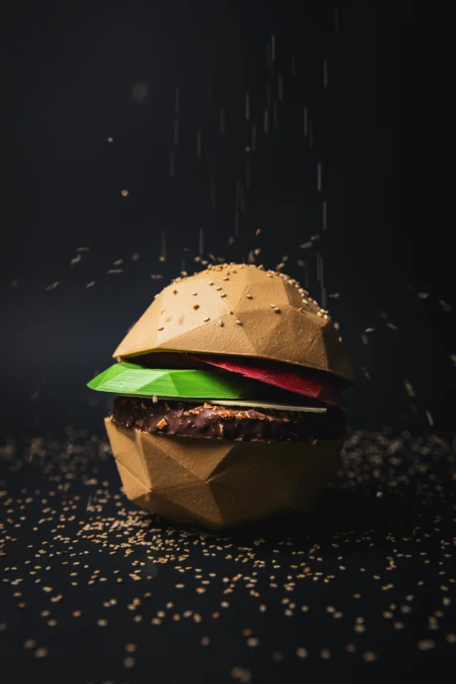 Mona de Pascua de bubó con forma de hamburguesa de chocolate, fotografiada sobre fondo negro con sésamo cayendo